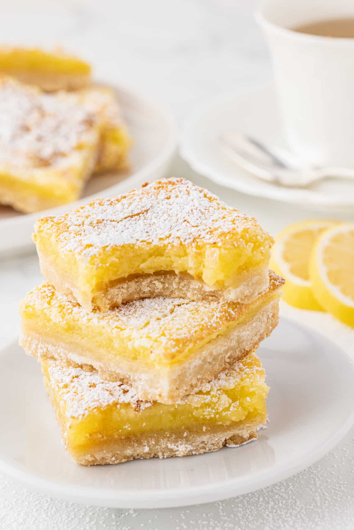 close up of stacked lemon bar squares on a white plate with a white tea up in the background