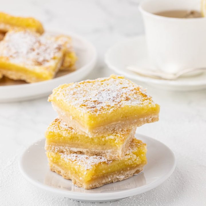 stack of lemon bars on a white plate and white marble countertop with a cup of tea in background
