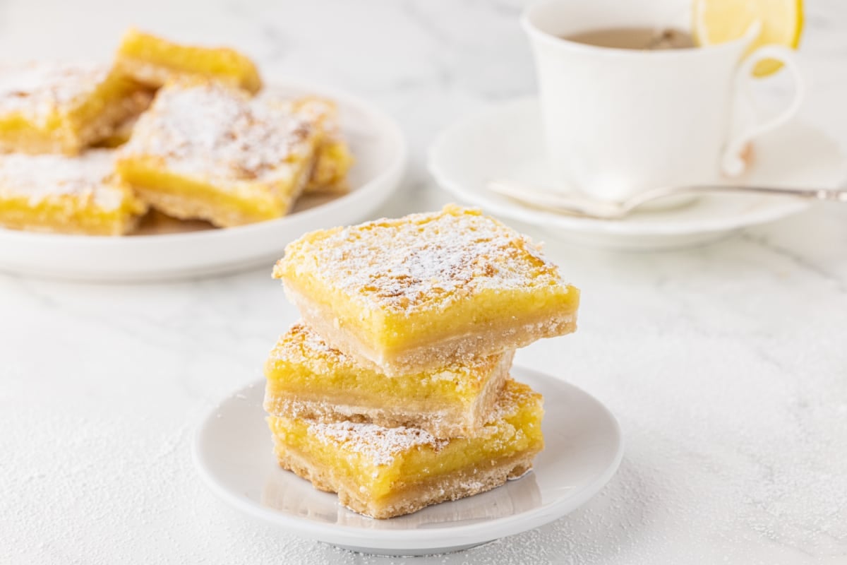 stack of lemon bars on a white plate and white marble countertop with a cup of tea in background