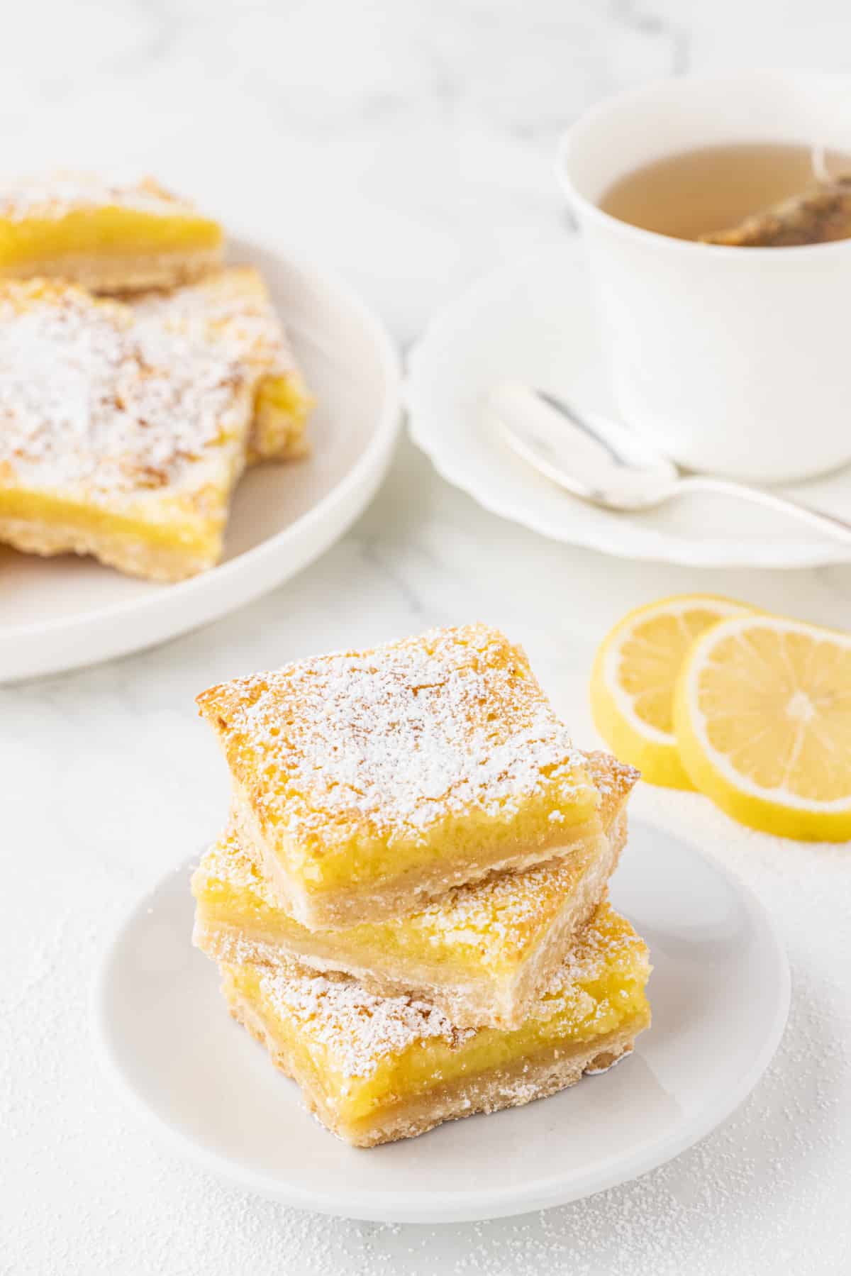 white plate with 3 lemon bars stacked by a cup of tea and lemon slices