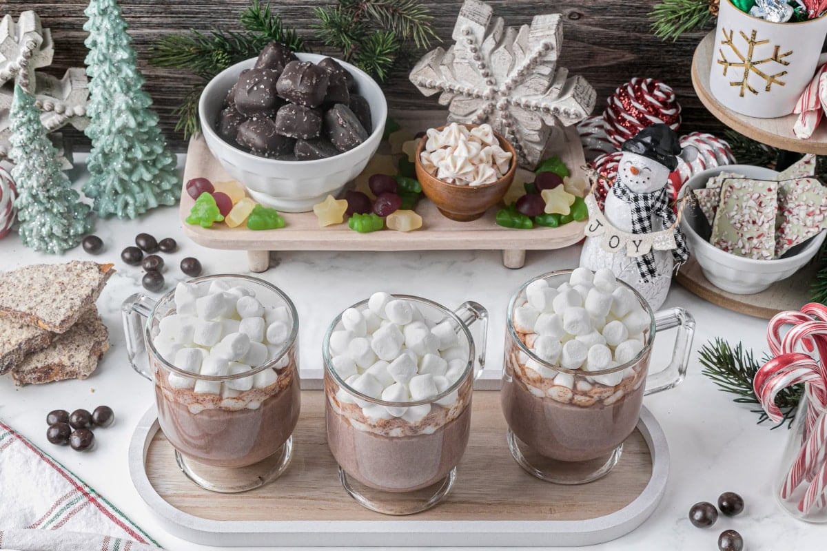 three cups of hot chocolate topped with marshmallows and on a tray with holiday decor in the background