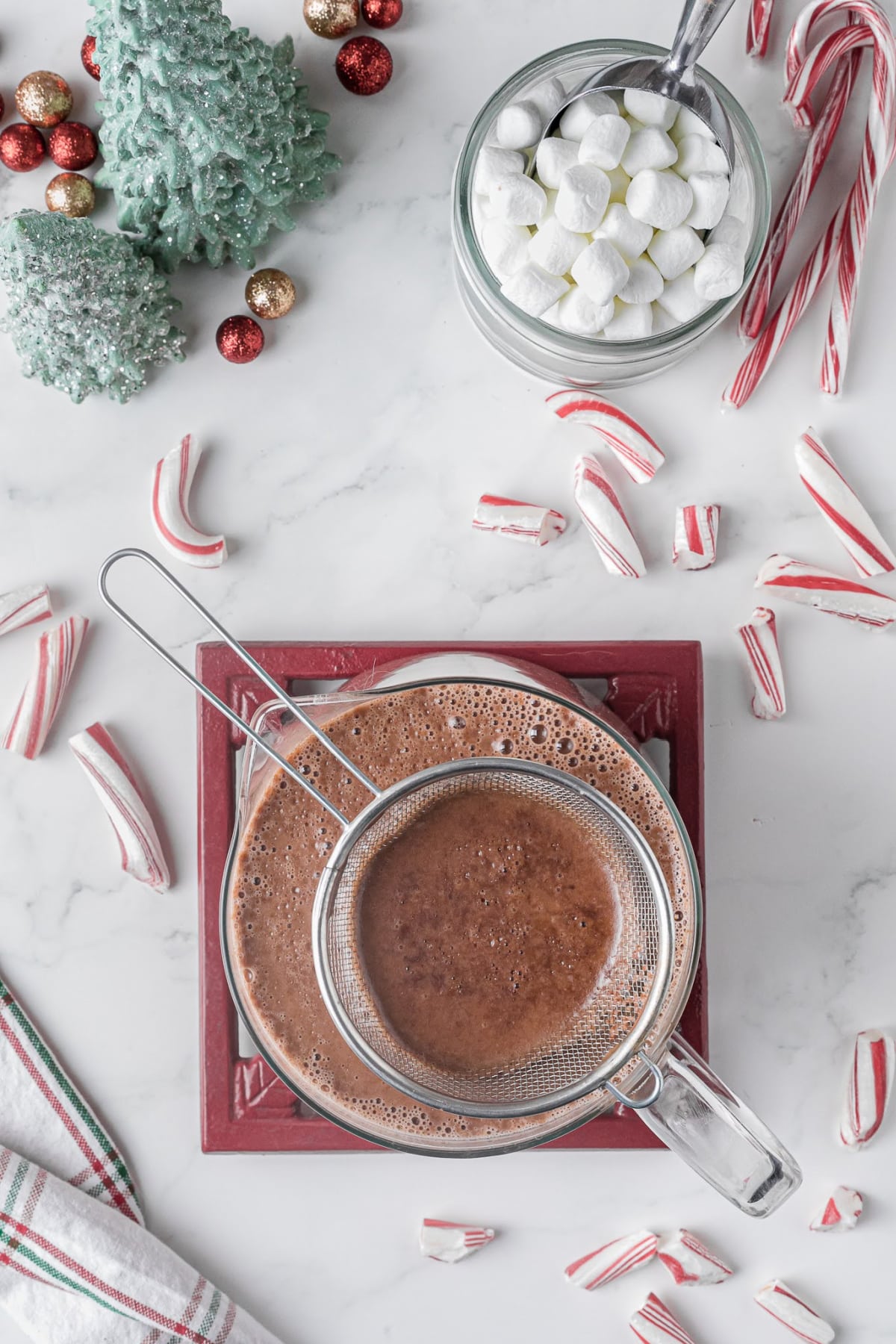 A strainer sitting in a large bowl of hot chocolate mix on a white countertop with candy canes and marshmallows