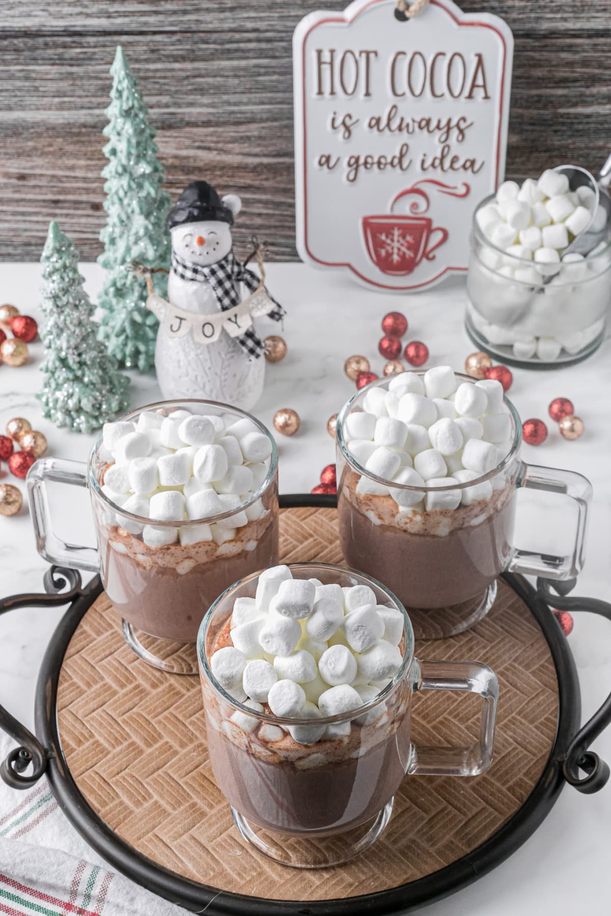 Three clear glass mugs of hot chocolate sitting on a table topped with marshmallows among holiday decorations