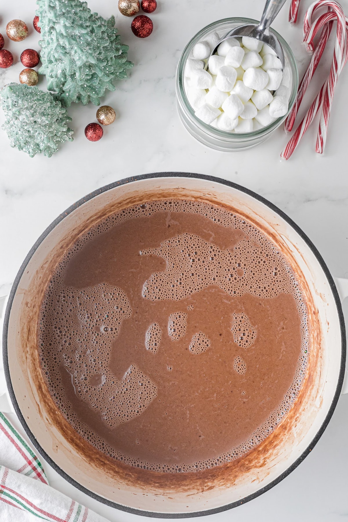 large saucepan full of hot chocolate on a white countertop with a cup full of marshmallows and holiday decor 