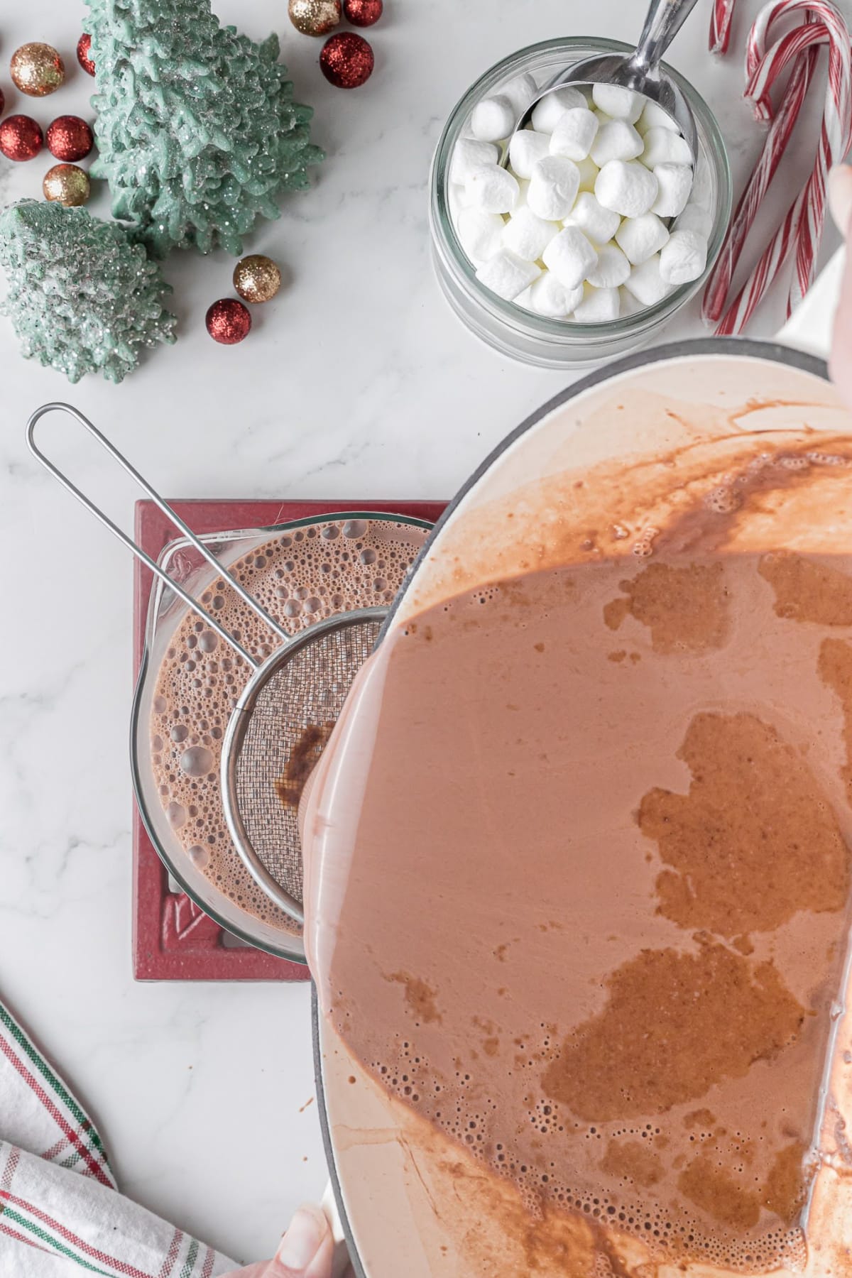 hot chocolate being strained through a strainer and into a bowl on a white counter top with a cup of marshmallows