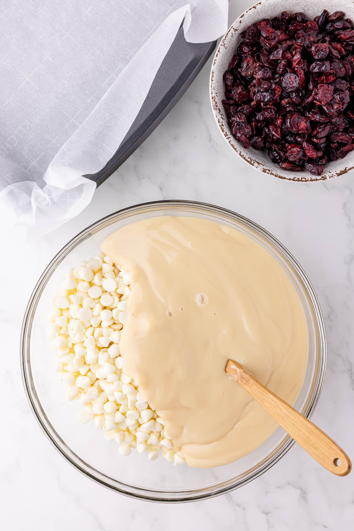 white chocolate chips melting in a glass bowl on a counter with a bowl of dried cranberries to the side