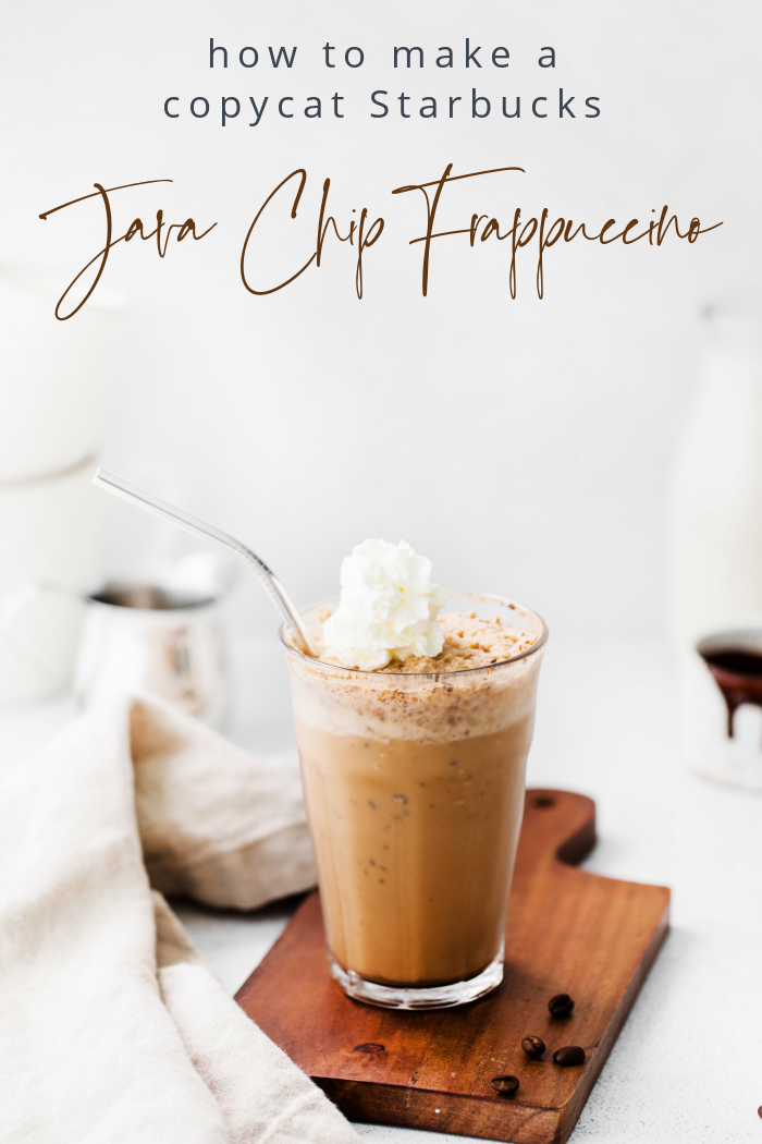 A large glass of homemade Java Chip Frappuccino sitting on a cutting board with coffee ingredients, whipped topping and coffee beans sprinkled around