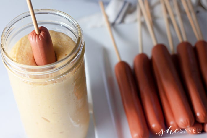 homemade corn dogs batter in a jar with a hotdog on a stick dipping in 