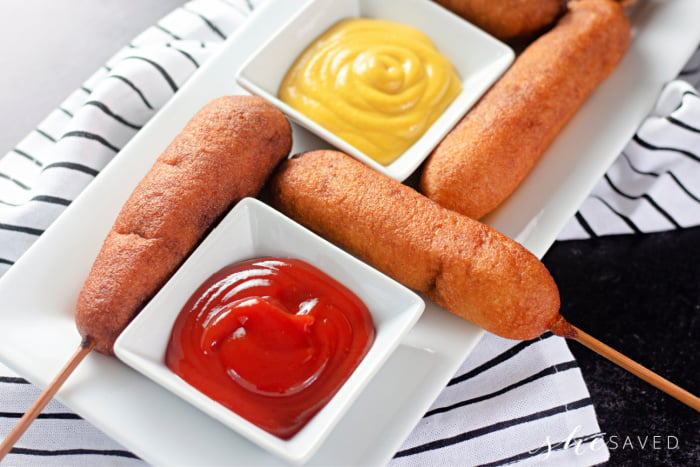 homemade corn dogs on a platter with condiments of ketchup and mustard