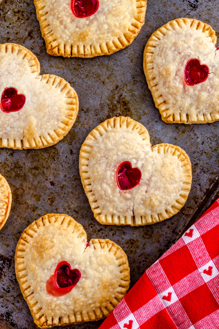 Mini strawberry filled heart shaped hand pies sitting on a cookie sheet with a red valentine linen