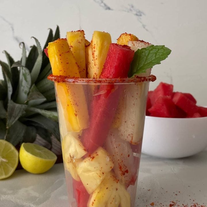A tall clear plastic cup full of fruit spears, a mint leaf and sprinkled with Tajin spice seasoning