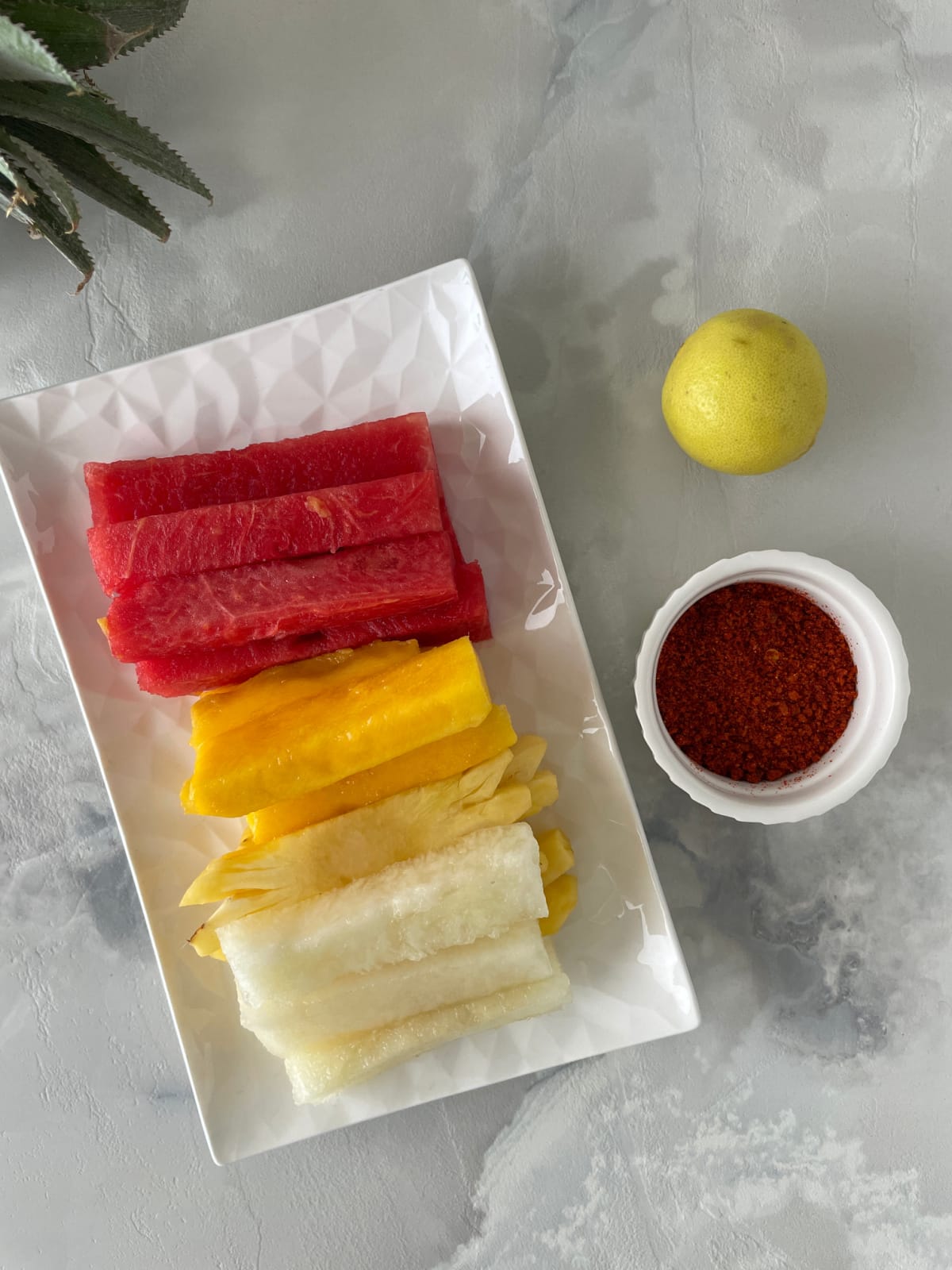 A white plate of watermelon, mango and jicama spears next to a bowl of chili seasoning and a lime